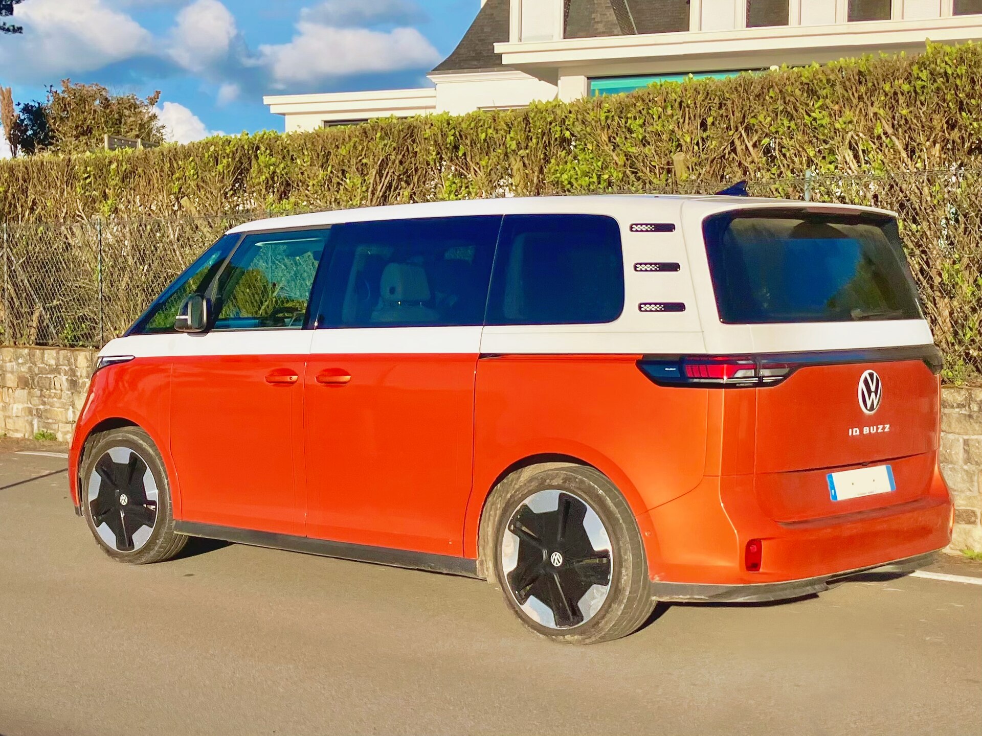 Orange och vit Volkswagen ID. Buzz parkerad vid en häck i kvällssol, sedd snett bakifrån