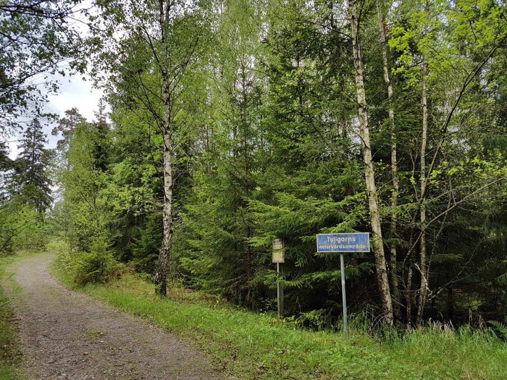 Blå skylt med texten Tullgarns naturvårdsområde vid en grusväg i blandskog