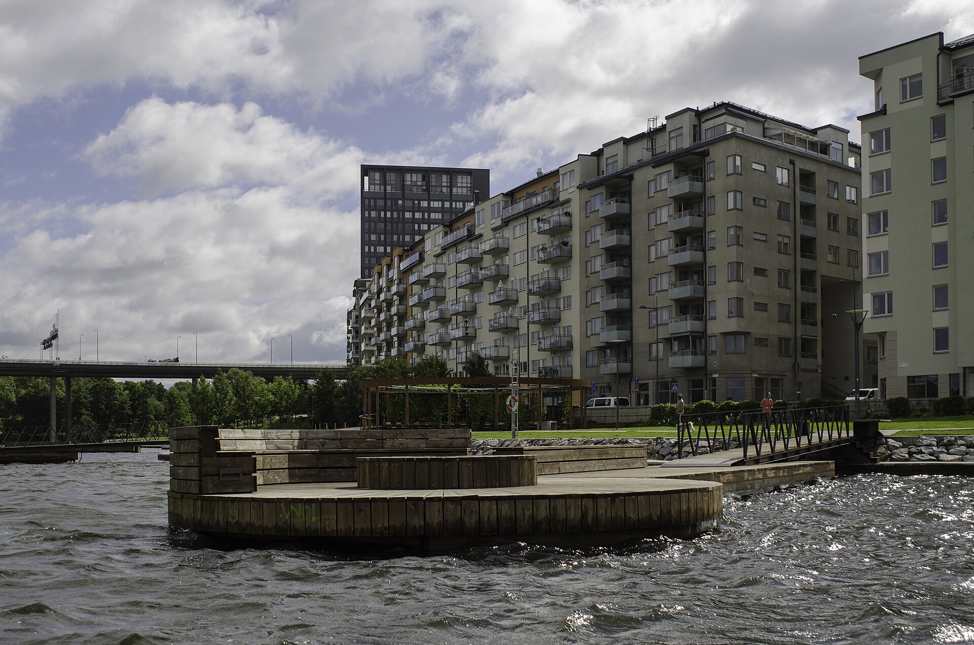 Träbrygga och pergola i Hornsbergs strandpark med flerbostadshus längs vattnet en molnig sommardag.