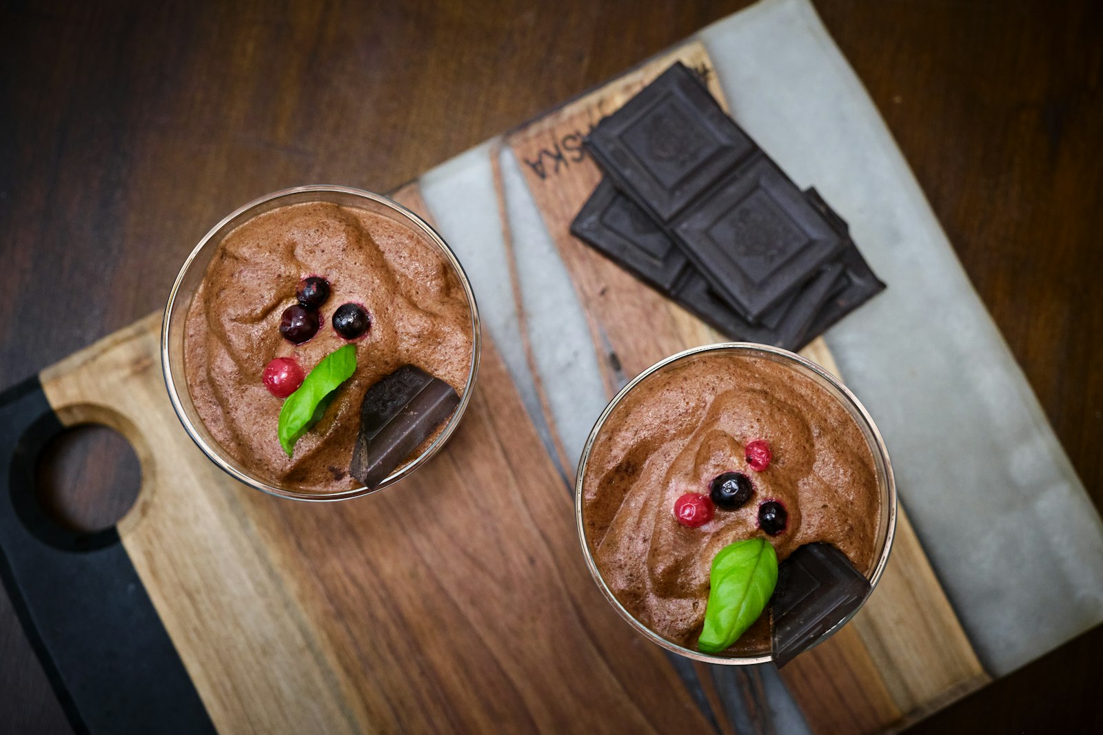 Två glas med luftig vegansk chokladmousse toppade med röda och svarta vinbär, ett grönt blad och bitar av mörk choklad