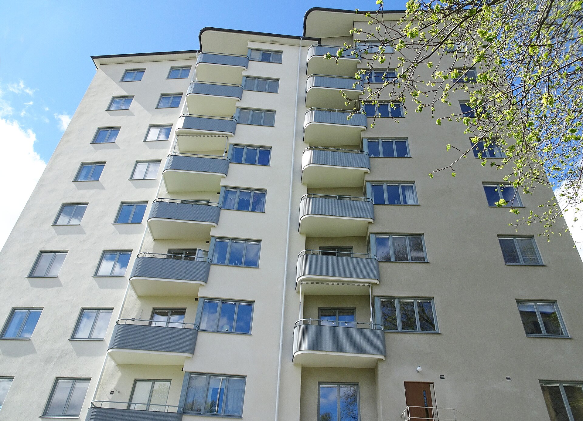 Funkishus i ljus puts på Pokalvägen på Reimersholme, med rundade balkonger i flera våningar mot blå himmel.