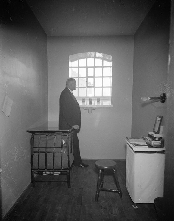 Fängelsecell på Långholmen med kalkade väggar, högt gallerförsett fönster och en man i kostym vid sängen