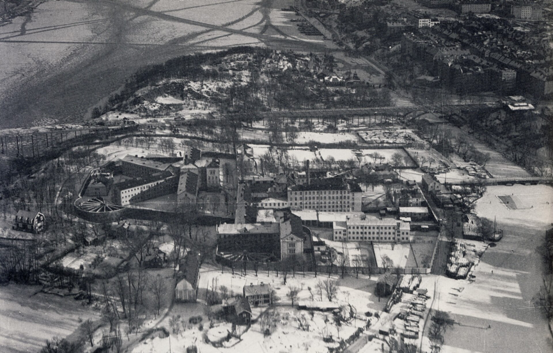 Snötäckt flygfoto över Långholmen med centralfängelsets flyglar, ringmuren och en solfjäderformad rastgård