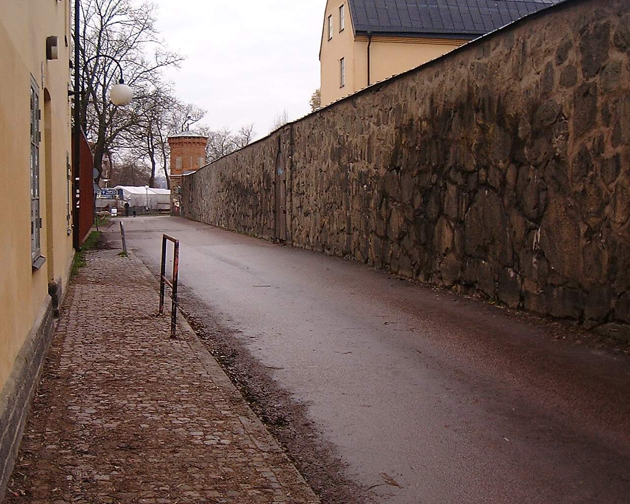 Hög gråstensmur längs en väg på Långholmen, med ett gult putsat hus till vänster och ett runt tegelvakttorn i fonden