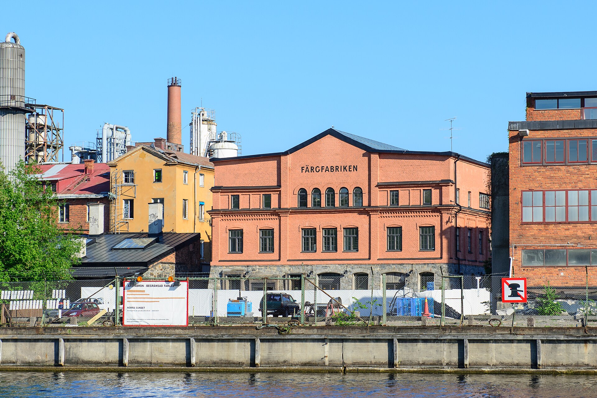 Färgfabrikens rosa tegelhus på Lövholmen med industrisilor och fabriksskorsten bakom, sett från vattnet