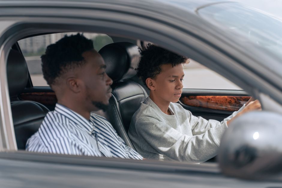 A father sits in the passenger seat while his teenage son practises driving, acting as the approved handledare.