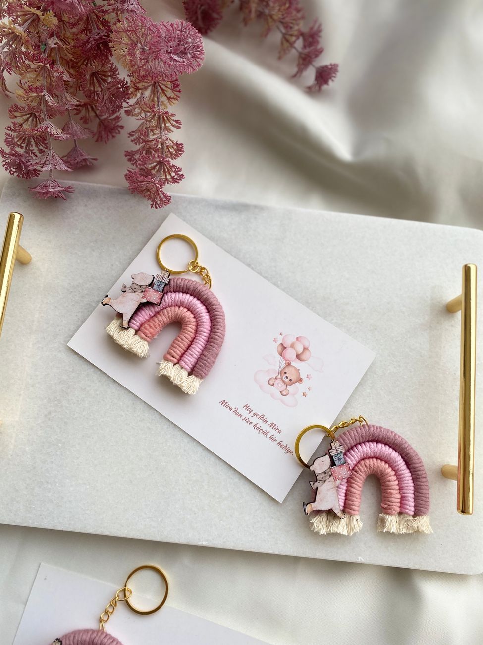 Två rosa nyckelringar av makramégarn formade som regnbågar, med fransar och guldfärgade ringar, på ett vitt kort