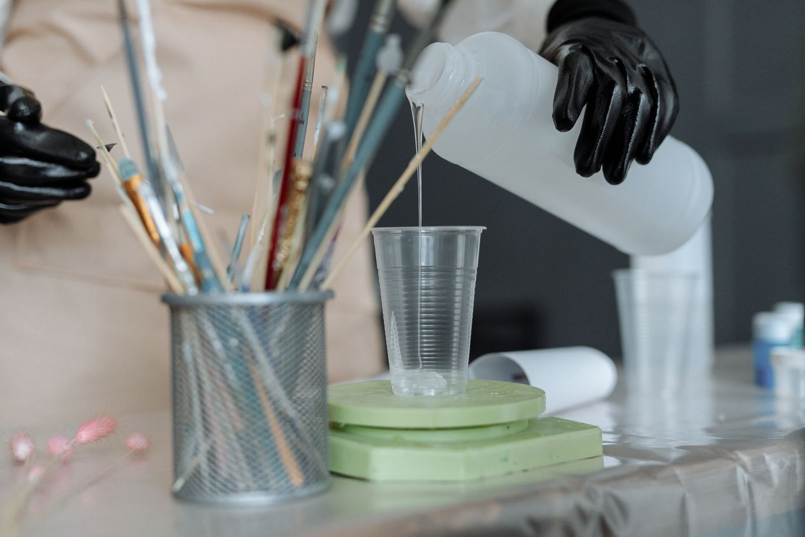Person med svarta skyddshandskar häller flytande resin ur en flaska i en genomskinlig plastmugg på ett arbetsbord