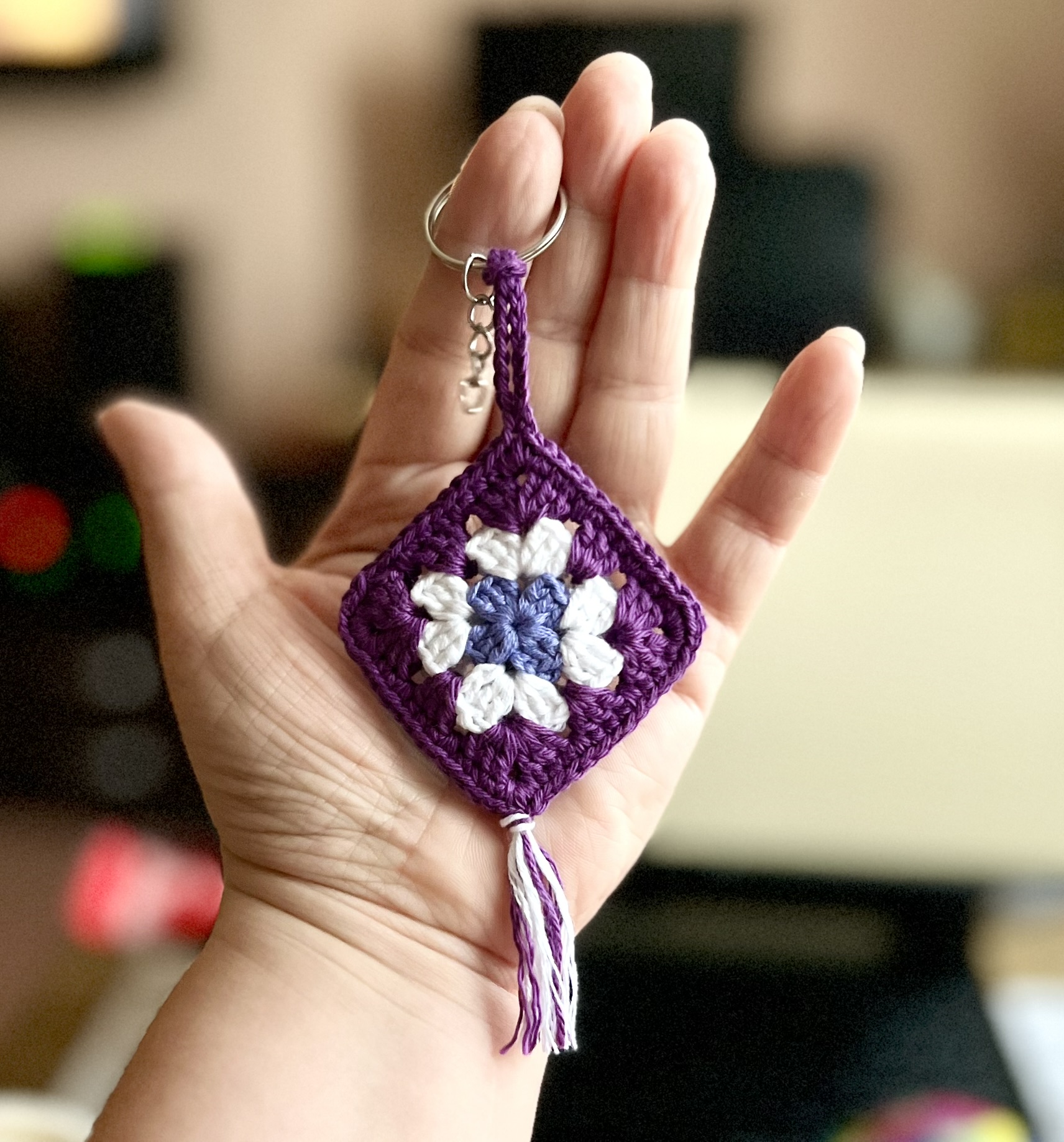 Virkad nyckelring i form av en granny square i lila, vitt och blått med tofs