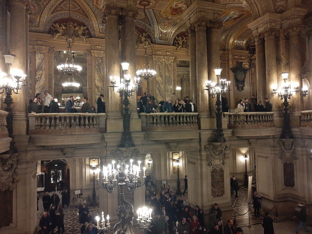 Den stora trappan i Palais Garnier med kandelabrar, marmorpelare och besökare på balkongerna