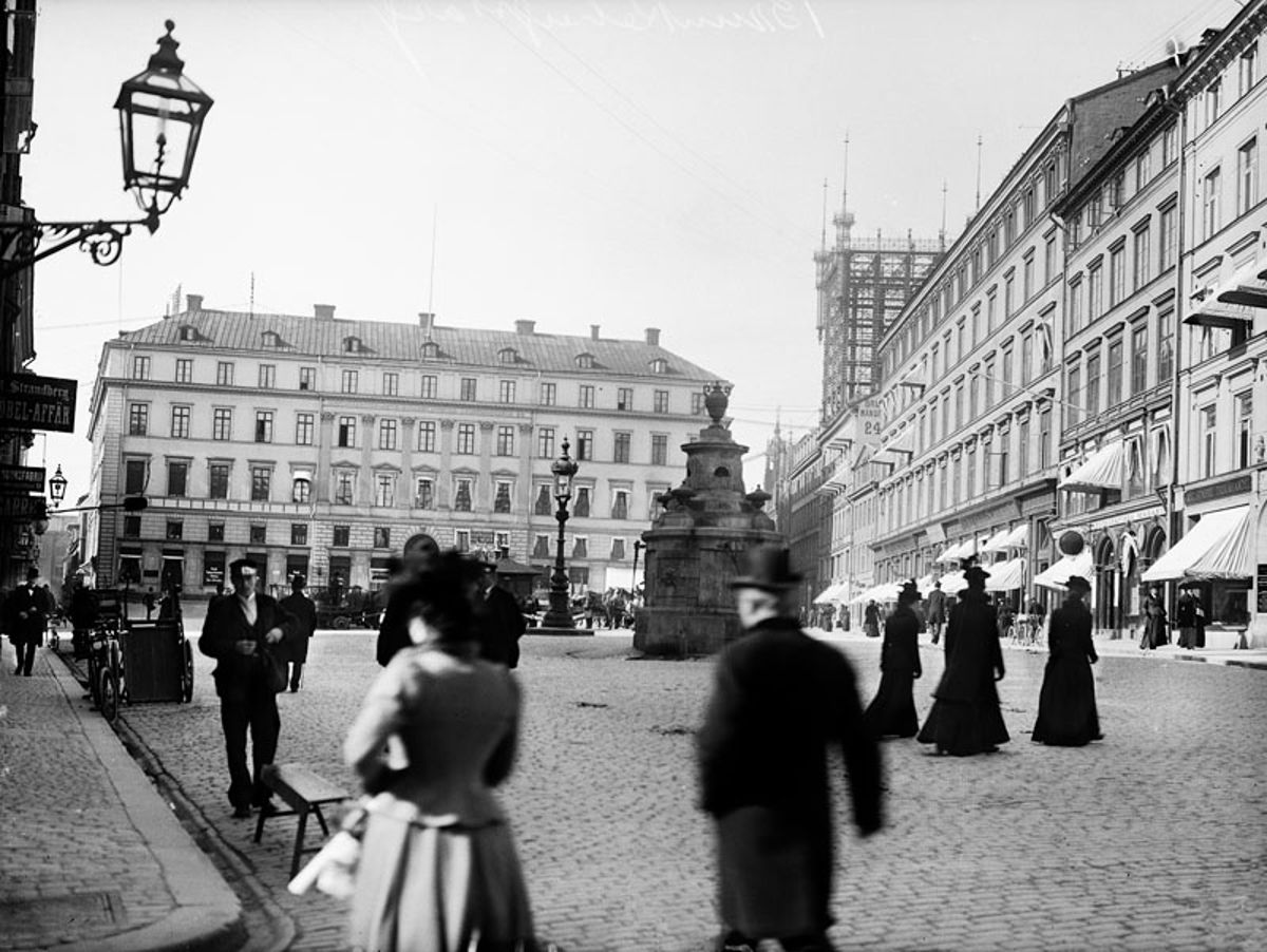 Kullerstensbelagta Brunkebergstorg 1899 med människor i hatt och långklänning, en rund stenbrunn mitt på torget och telefontornets järnställning bakom husraden