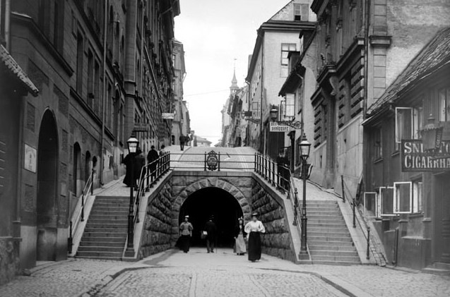 Svartvitt fotografi från 1896 av en välvd tunnelmynning i sten, med trappor på båda sidor upp till en gata ovanför och fotgängare i långa kjolar framför öppningen