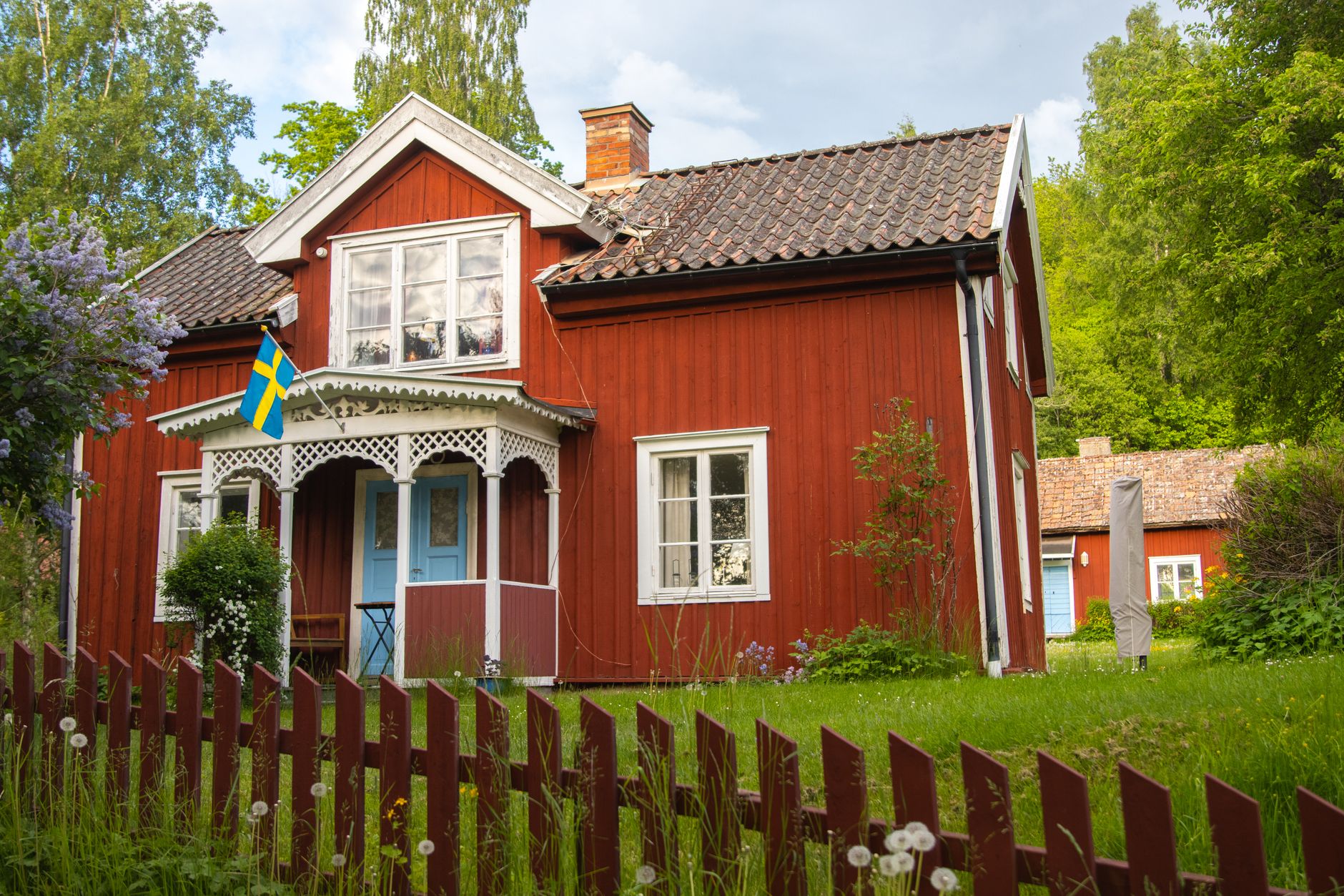 Rött trähus med vit veranda och svensk flagga bakom ett rött staket.