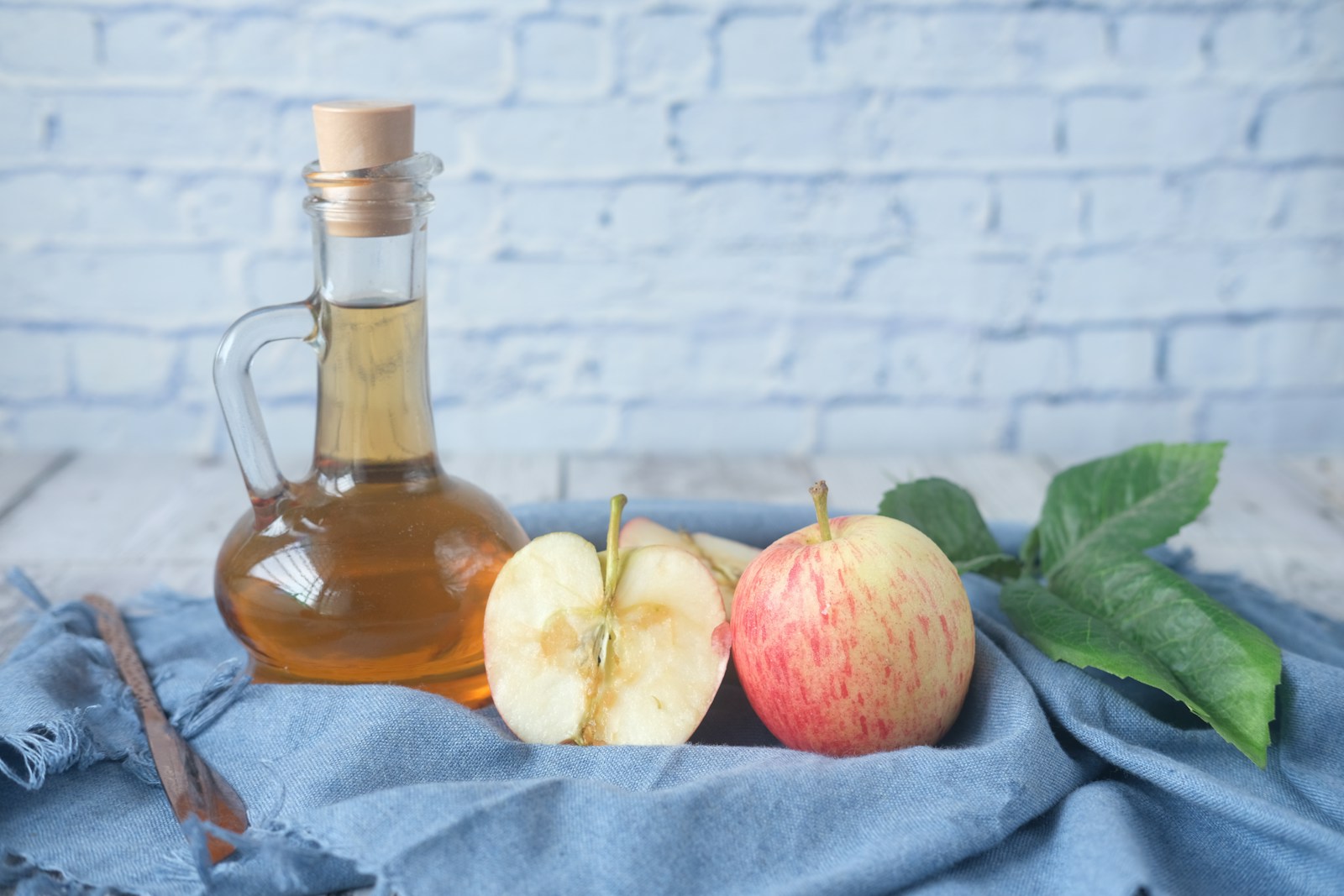 Vinägerflaska av glas bredvid halverat och helt äpple på en blå linneduk