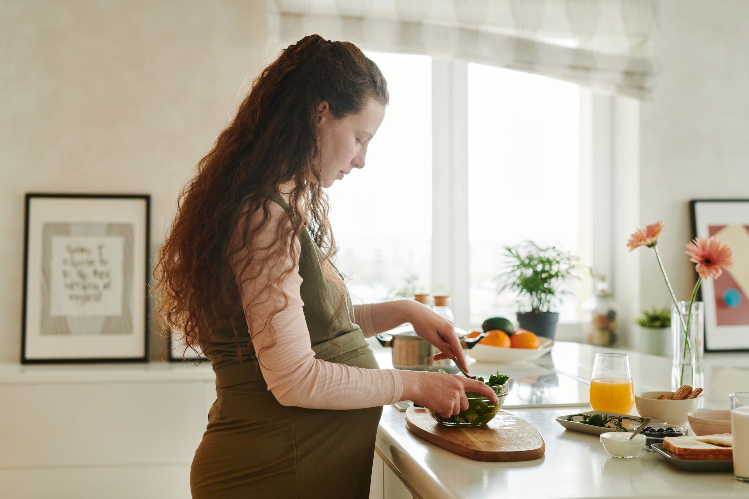 Gravid kvinna skär grönsaker vid en köksbänk med frukt och fullkornsbröd bredvid