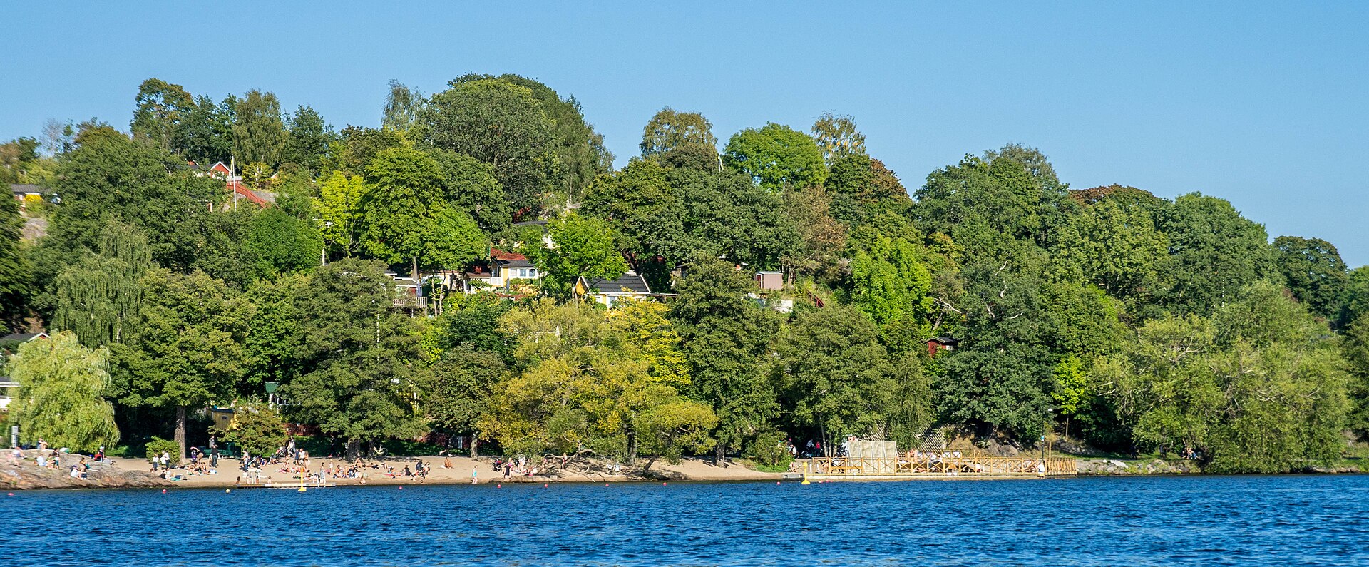 Tanto strandbad sett från Årstaviken med badande och grönskande strandsluttning