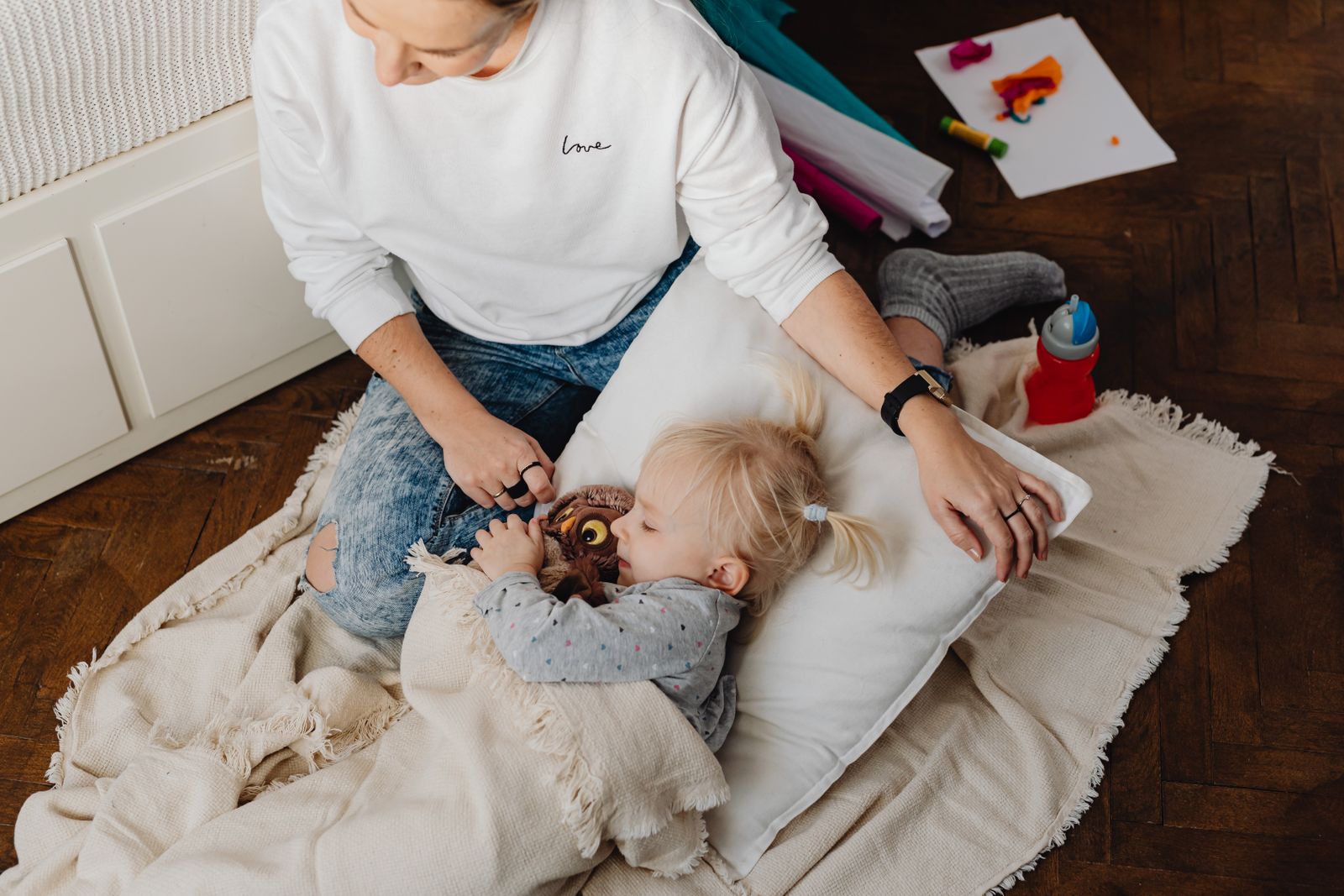 A parent sitting on the floor beside a small child asleep on a pillow with a soft toy