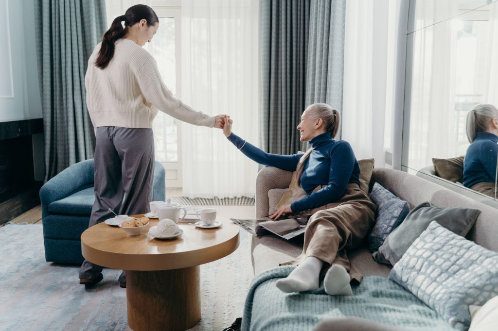 Vuxen dotter håller sin äldre mammas hand i ett ljust vardagsrum