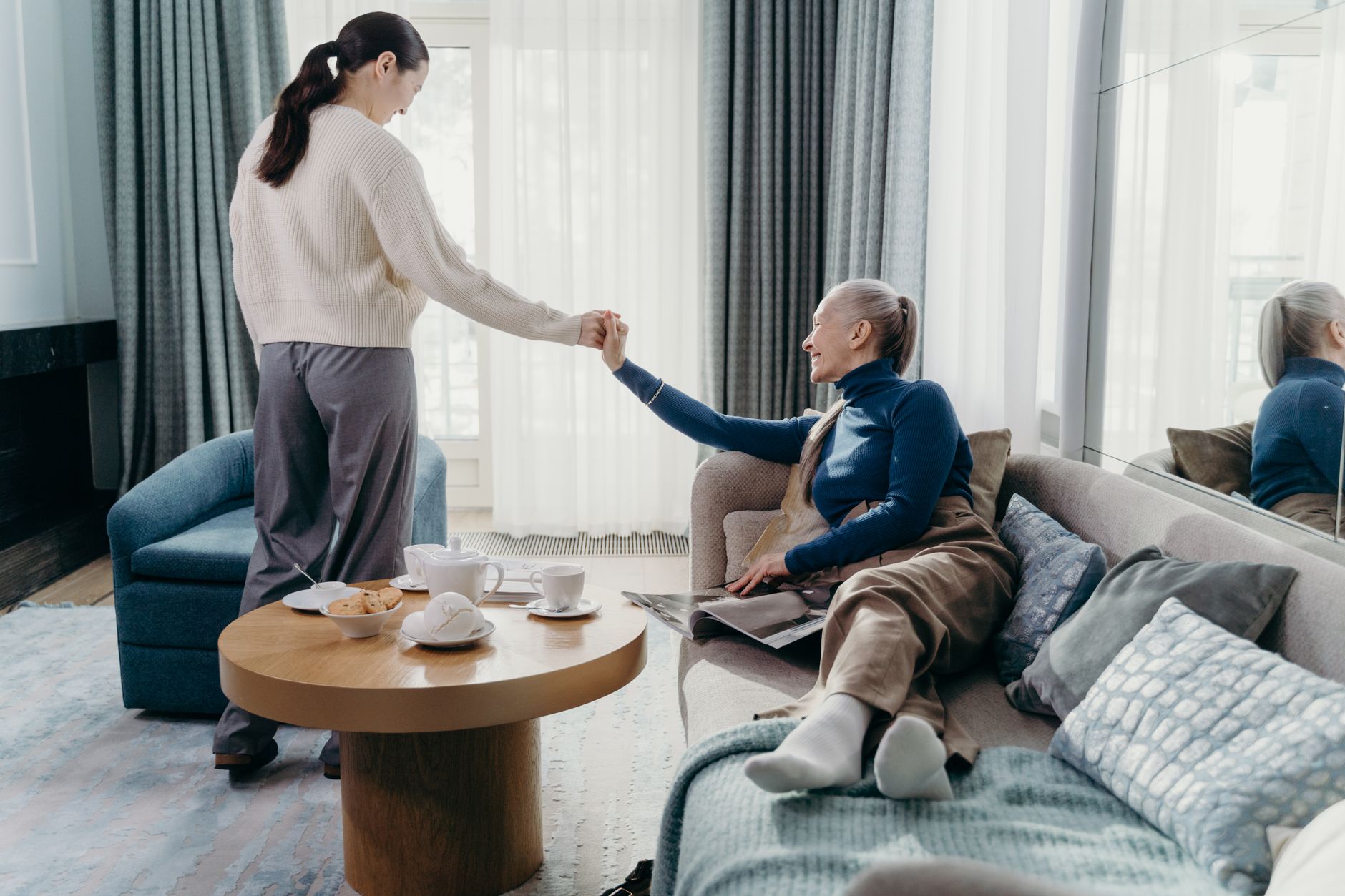 Vuxen dotter håller sin äldre mammas hand i ett ljust vardagsrum