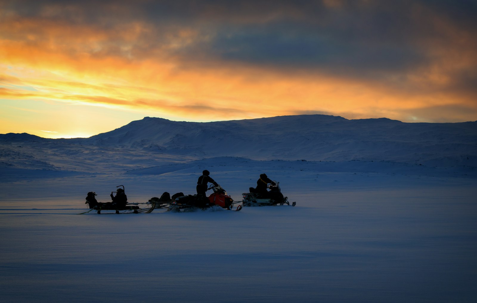Två snöskotrar på snötäckt fjäll i solnedgång, den ena drar en släde