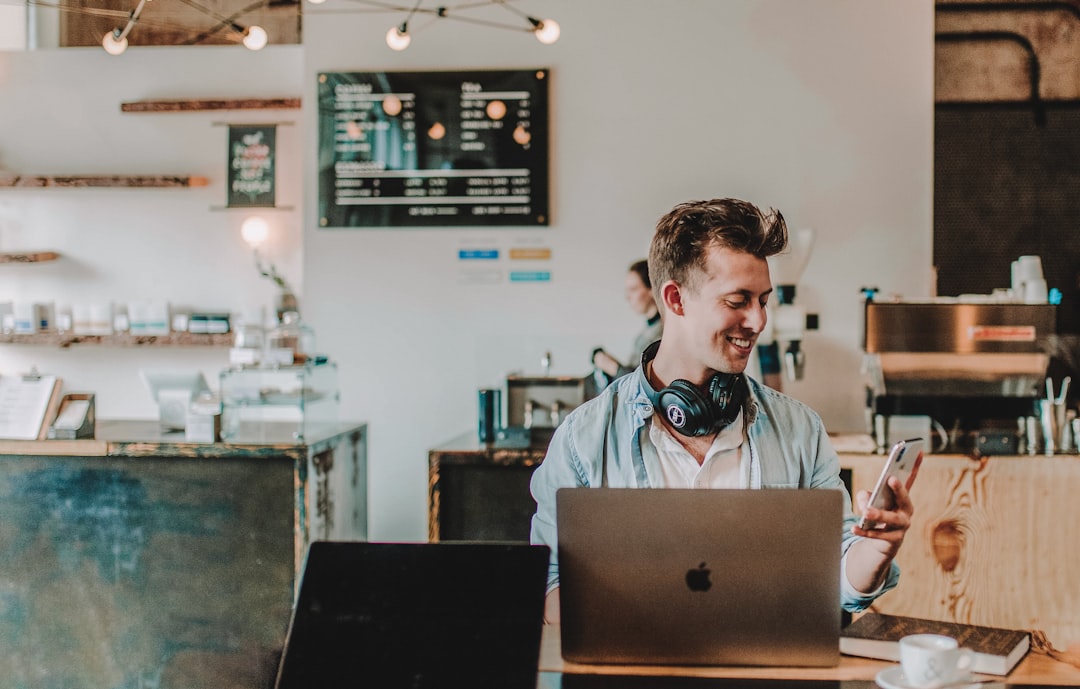 Leende person vid en laptop som tittar på sin telefon på ett kafé