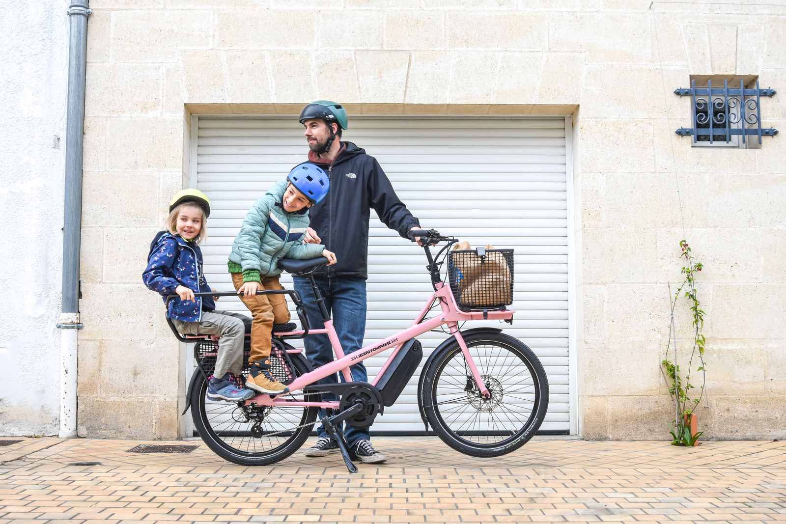 Pappa med två barn i hjälm vid en rosa lastcykel med barnbänk över bakhjulet