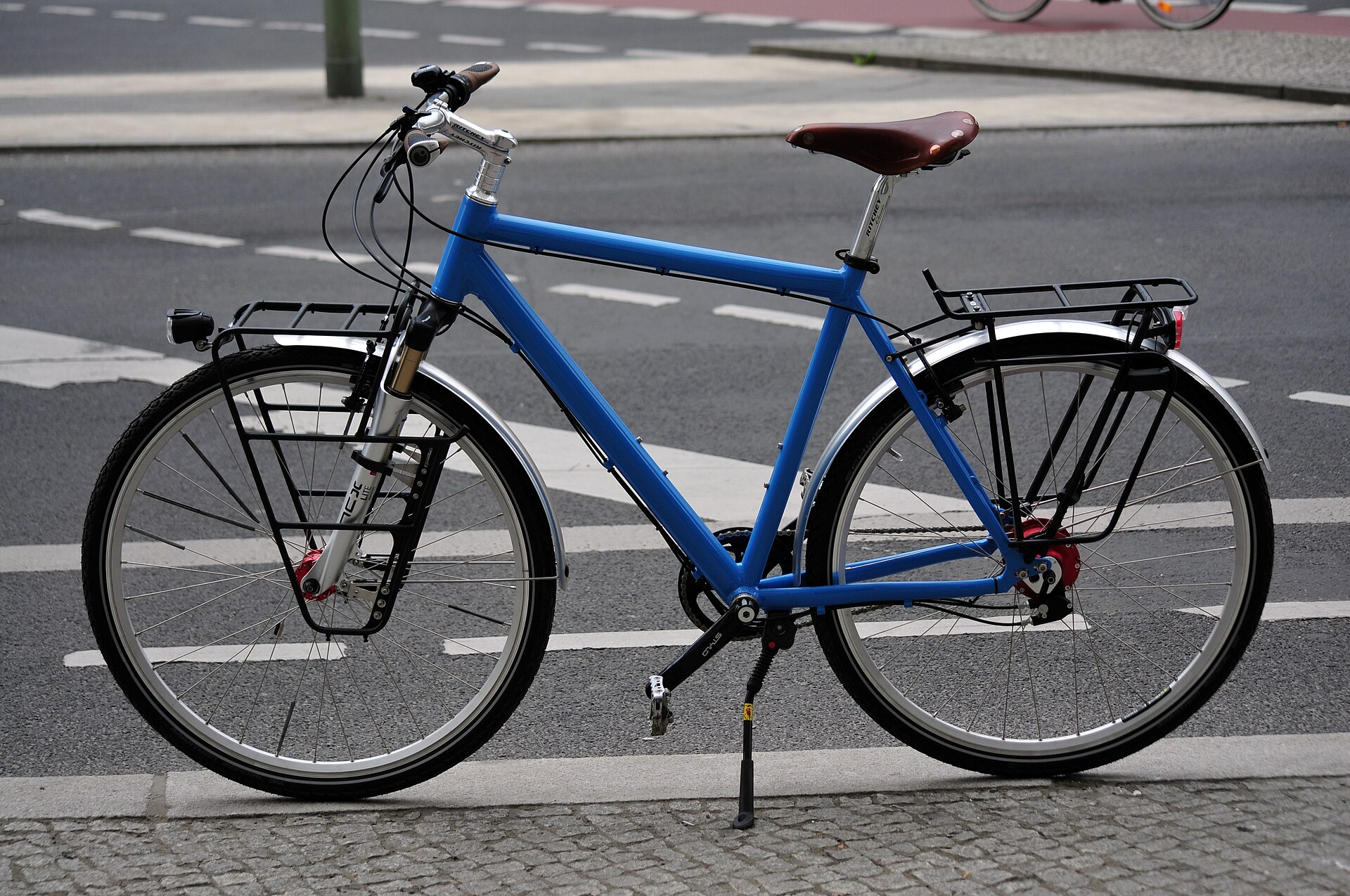 Blå trekkingcykel i profil med stänkskärmar, pakethållare bak, lågt monterad hållare fram, navdynamo och stöd