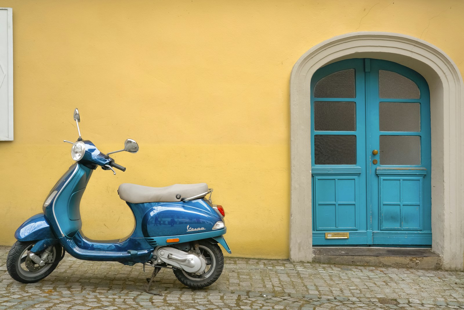Blå moped parkerad på kullerstenar framför en gul husvägg med turkos dörr
