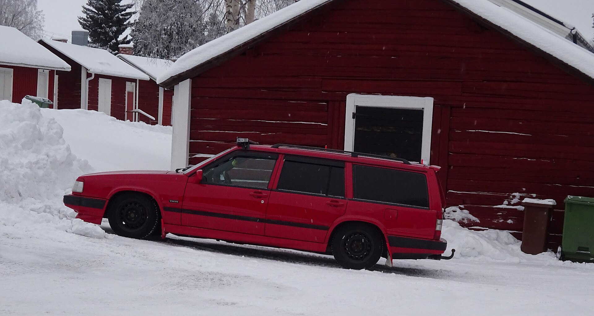 Röd ombyggd Volvo kombi i snön framför röda timmerhus