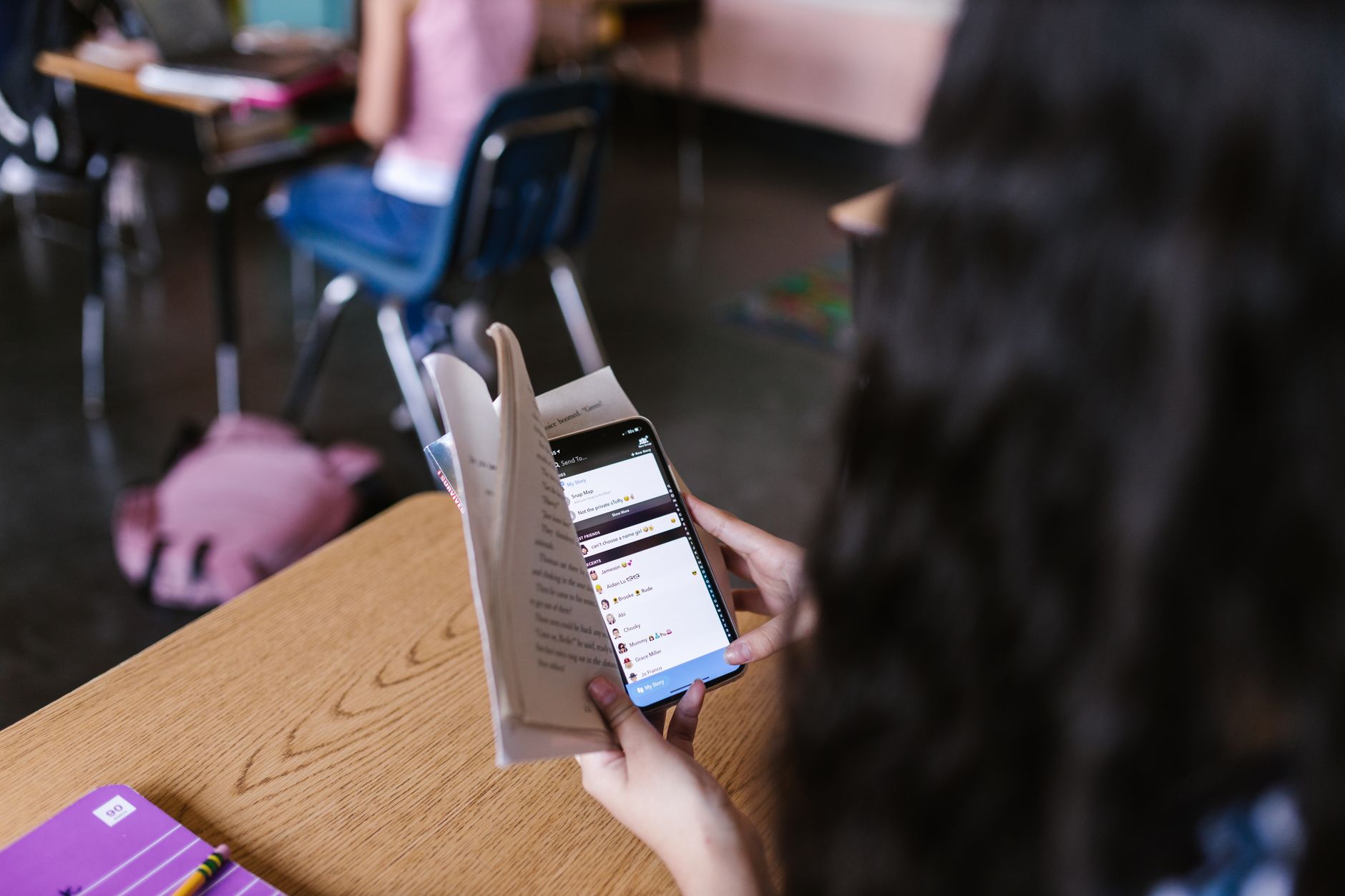 Student i klassrum håller mobilen bakom en uppslagen bok och läser en chatt i stället för texten