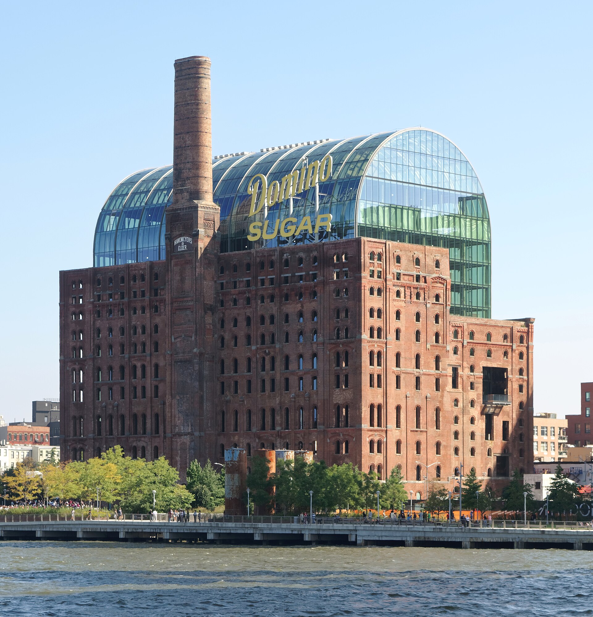 Det gamla sockerraffinaderiet Domino i tegel med skorsten, glaskupol och gul Domino Sugar-skylt, sett från East River med Domino Park längs stranden