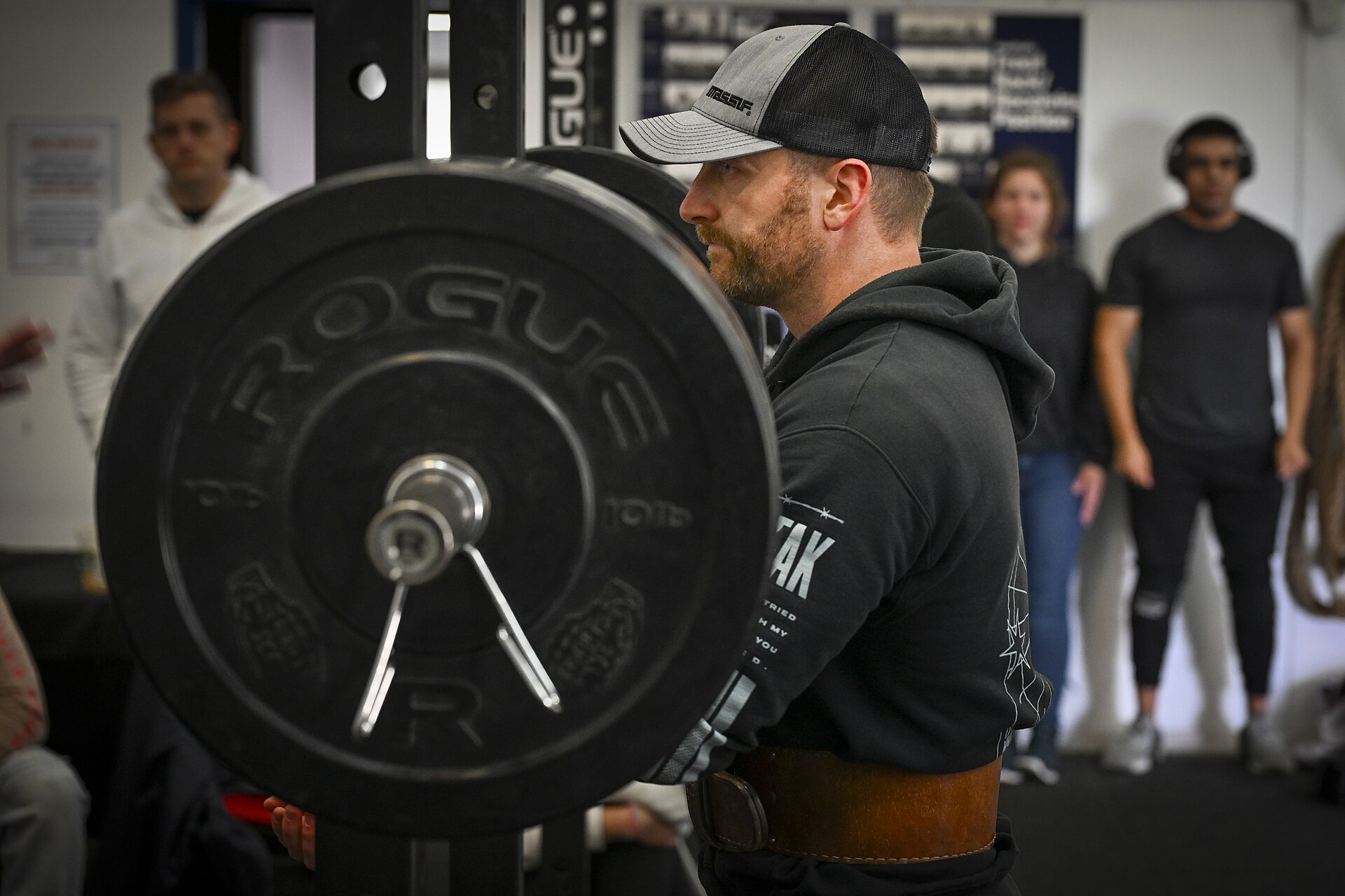 En man med bälte och keps ställer sig vid en lastad skivstång i ett knäböjsställ medan åskådare tittar på i ett gym