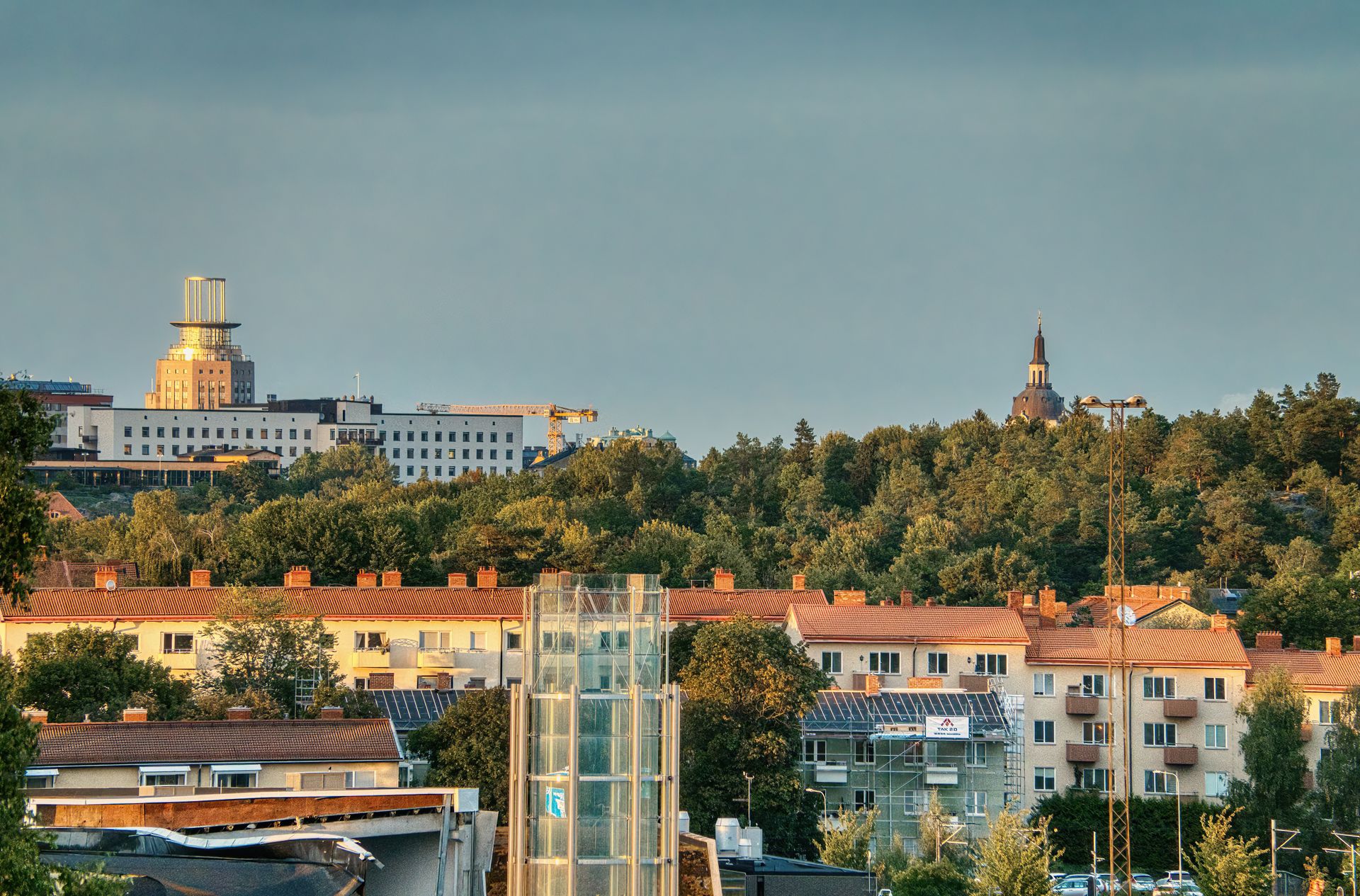 Flerbostadshus med tegeltak framför en skogsklädd höjd i Stockholm, med innerstadens siluett i bakgrunden