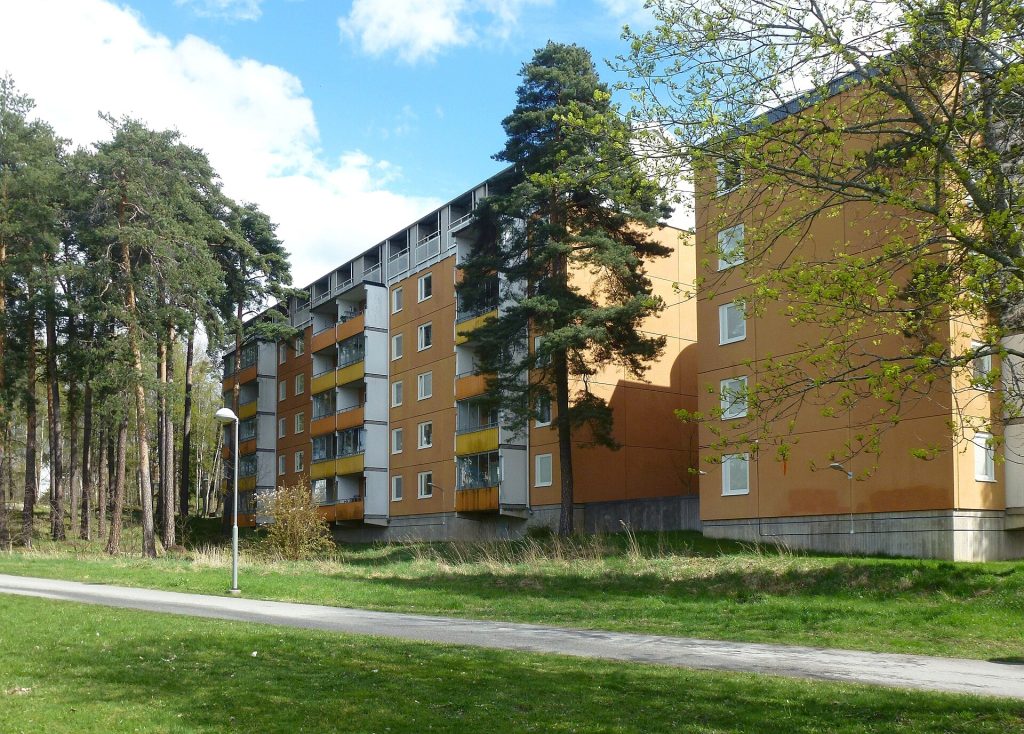Bostadshus i orange och gult från 1970-talet i Husby, med tallskog och gångväg framför