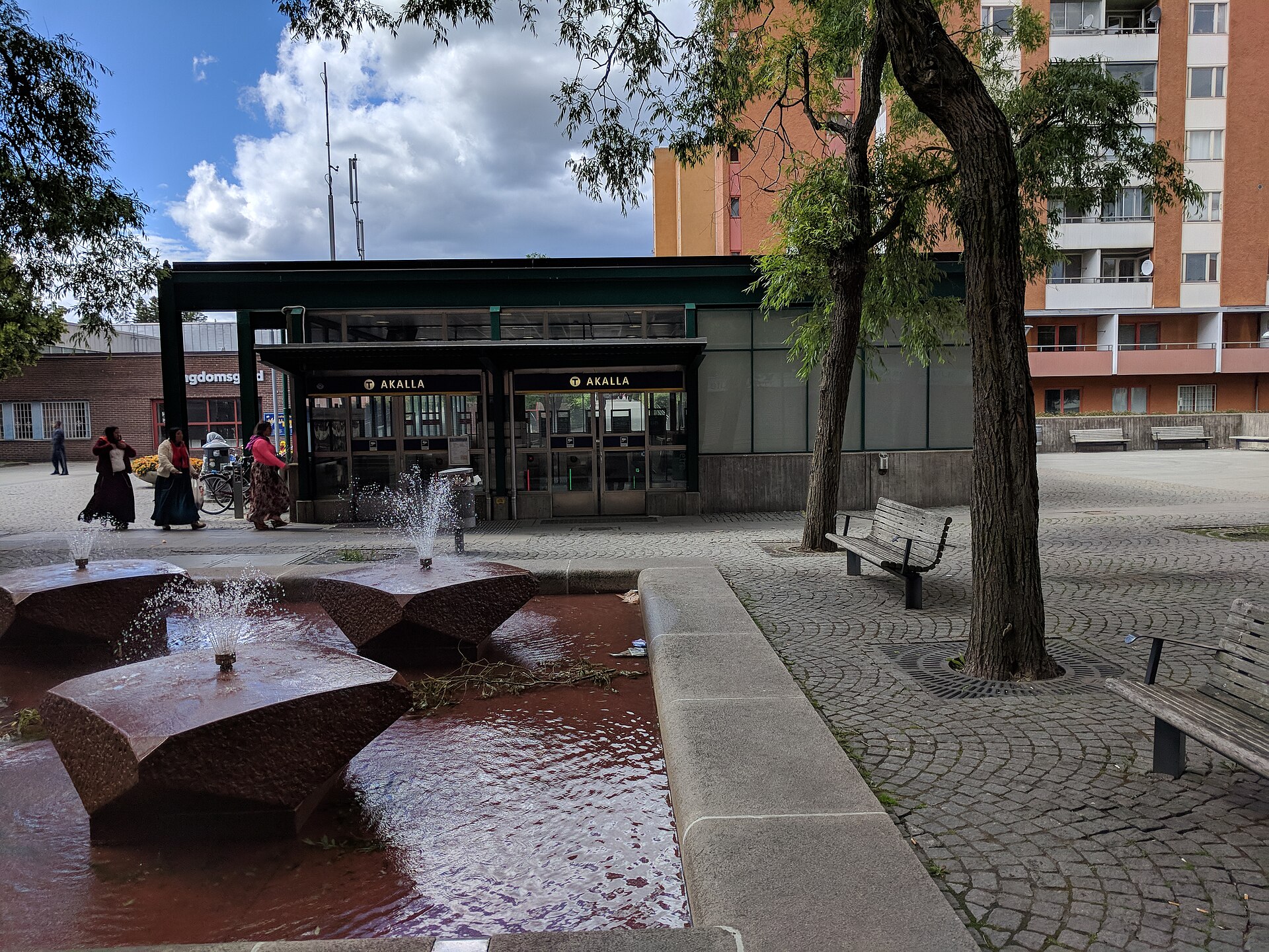 Den glasade uppgången till Akalla tunnelbanestation med en fontän i röd sten och bänkar framför
