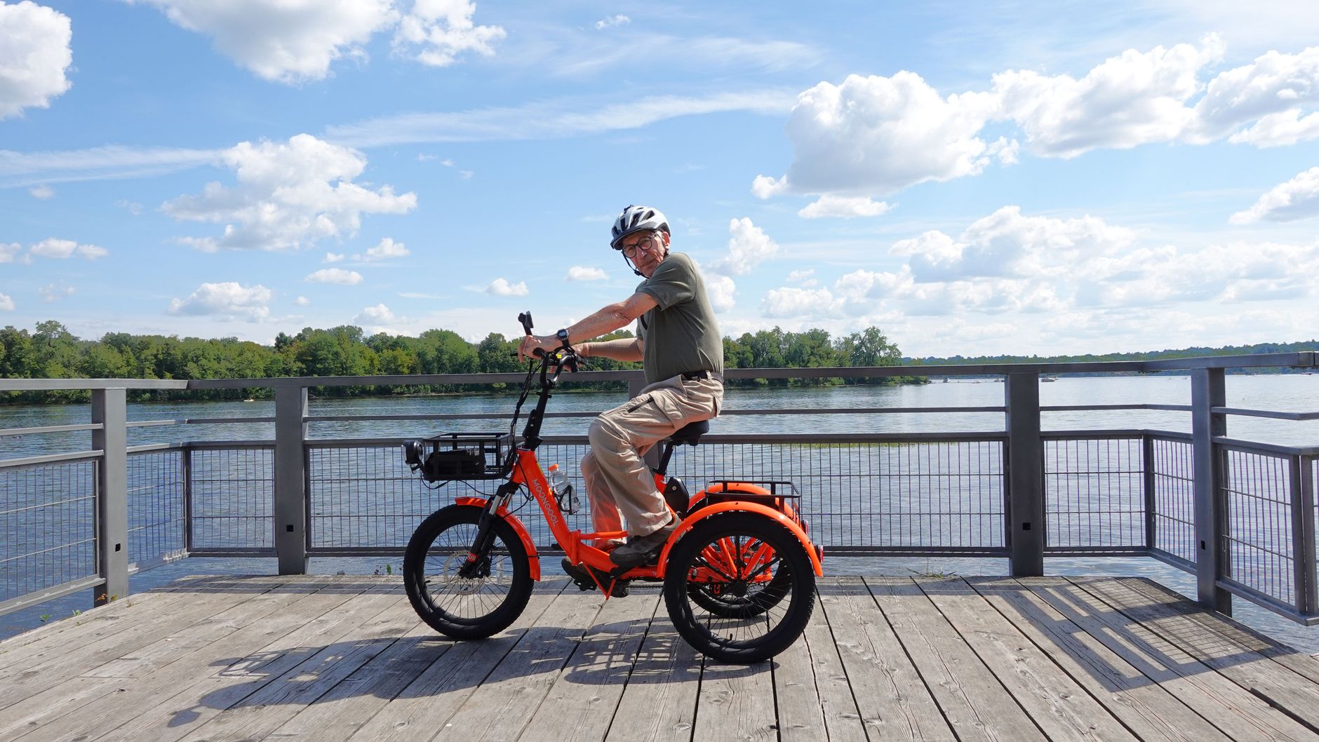 Äldre man med hjälm på en orange trehjulig elcykel vid en sjö