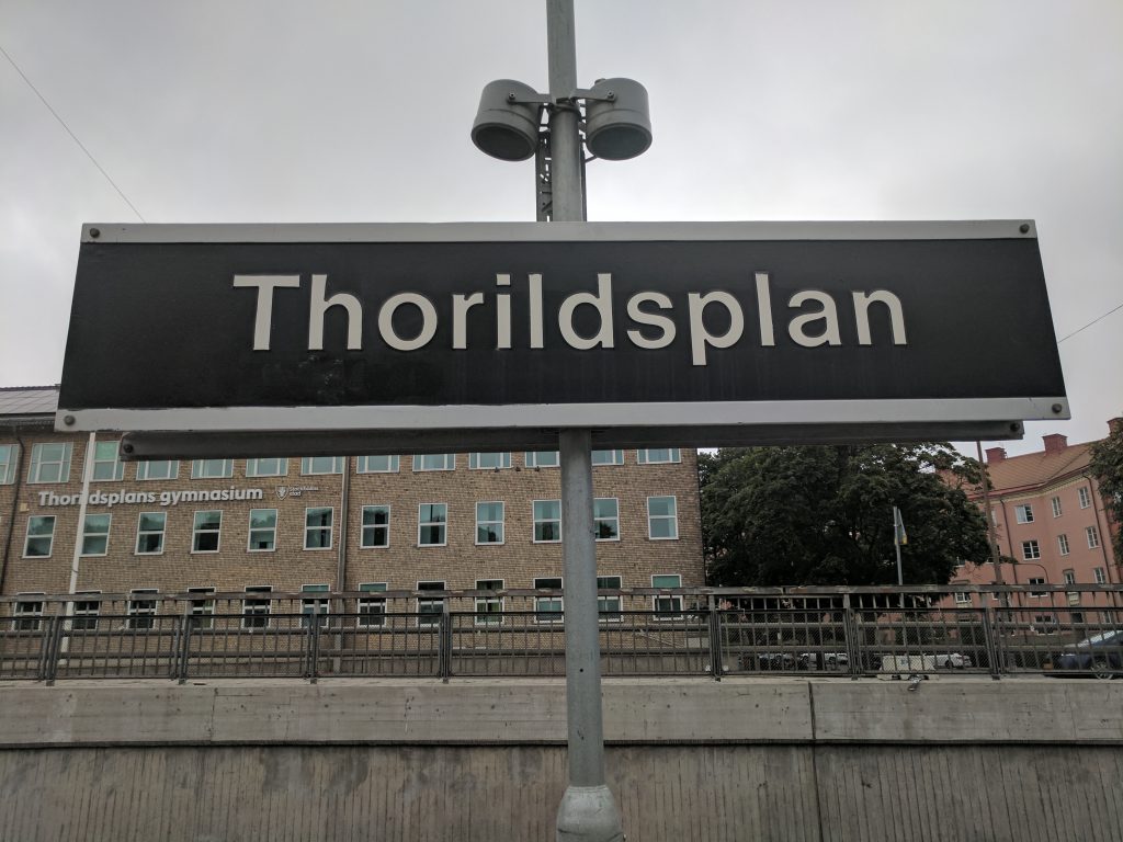 Stationsskylten Thorildsplan med Thorildsplans gymnasium i gult tegel bakom