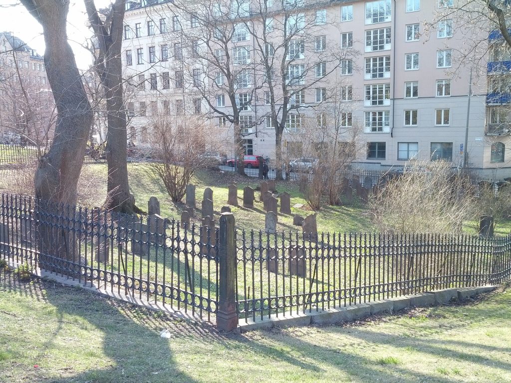 Gjutjärnsstaketet runt Judiska begravningsplatsen Kronoberg med gravstenar innanför och bostadshus i bakgrunden.