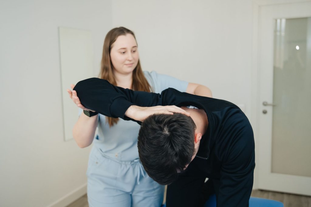 Fysioterapeut som stretchar ryggen och armen på en framåtböjd patient i ett ljust behandlingsrum.