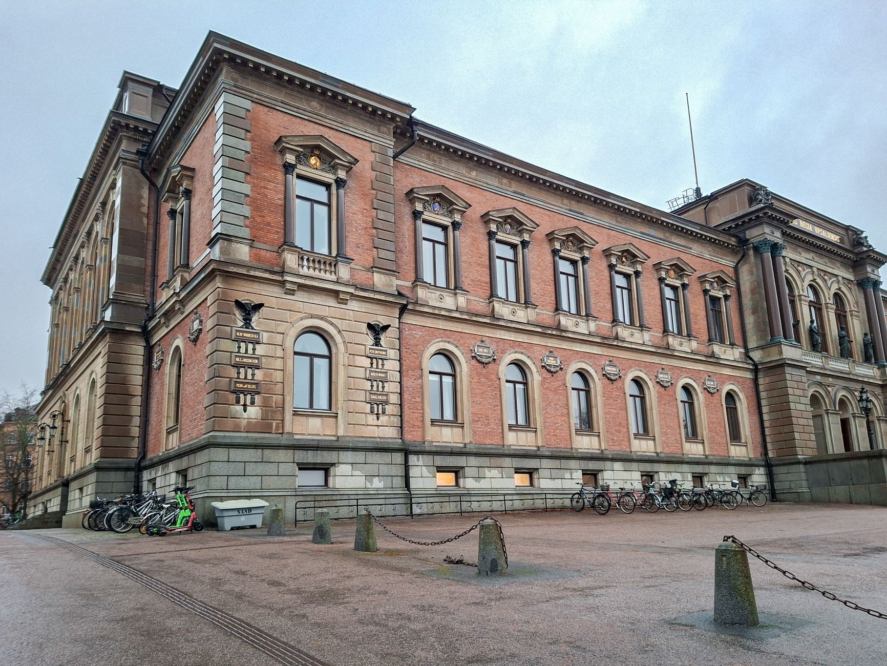Universitetshuset i Uppsala i rött tegel och ljus sten, med cyklar längs fasaden och namnplattor över tidigare professorer