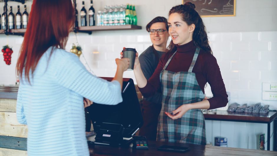 Ung person arbetar som barista och lämnar över en kaffe över disken i ett kafé