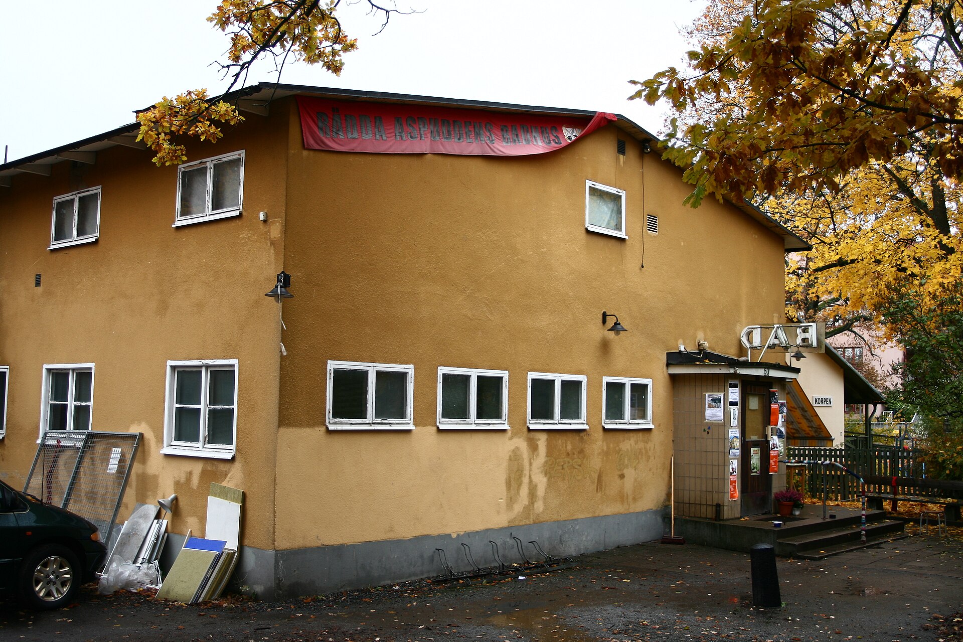 Det gula badhuset i Aspudden med en röd banderoll med texten Rädda Aspuddens badhus under takfoten och en BAD-skylt över entrén