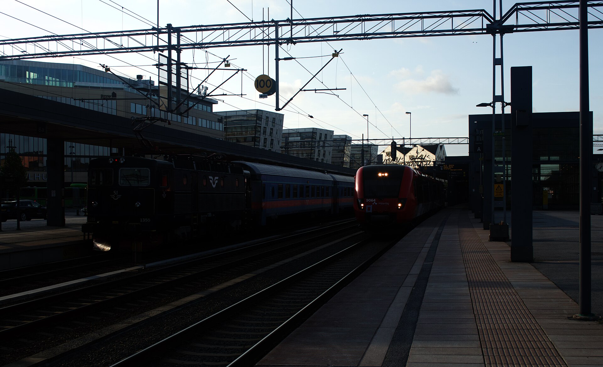 Två tåg står vid plattformarna på Uppsala centralstation i motljus, med kontaktledningar och stationsbyggnader i bakgrunden