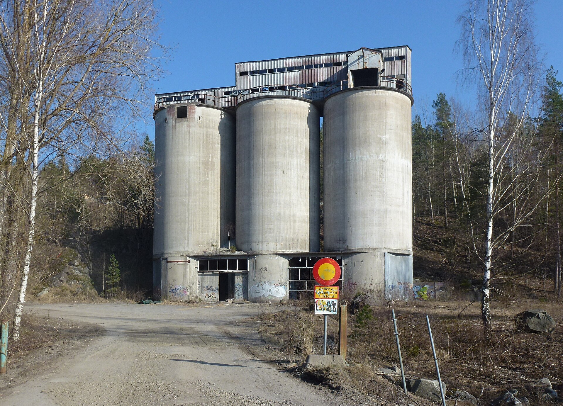 Höga kalkstenssilor i betong vid den nedlagda cementfabriken i Stora Vika, sedda från grusvägen fram till anläggningen