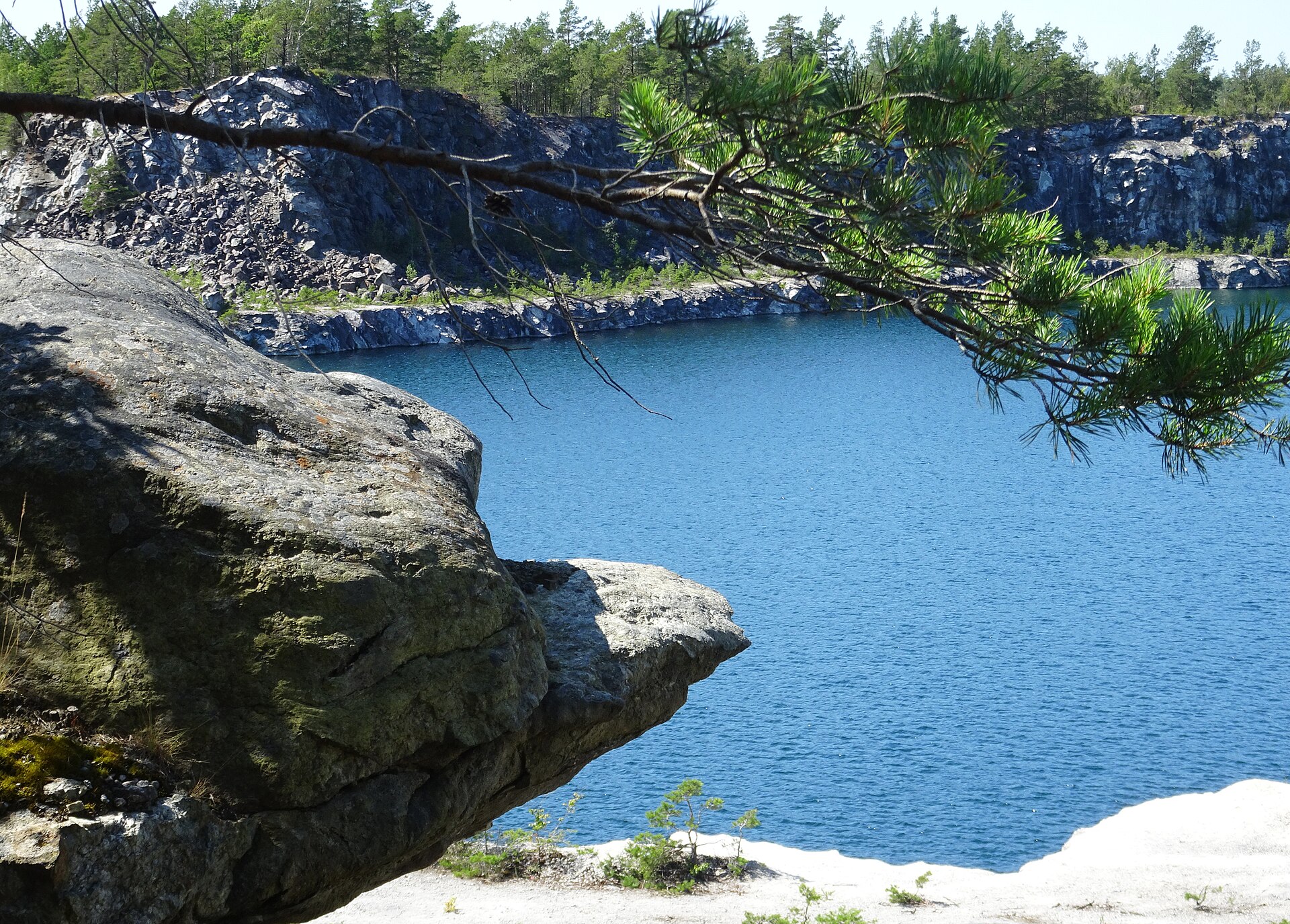 Den djupblå brottsjön Sänket i Stora Vika kalkbrott med en mörk bergklippa och en tallgren i förgrunden