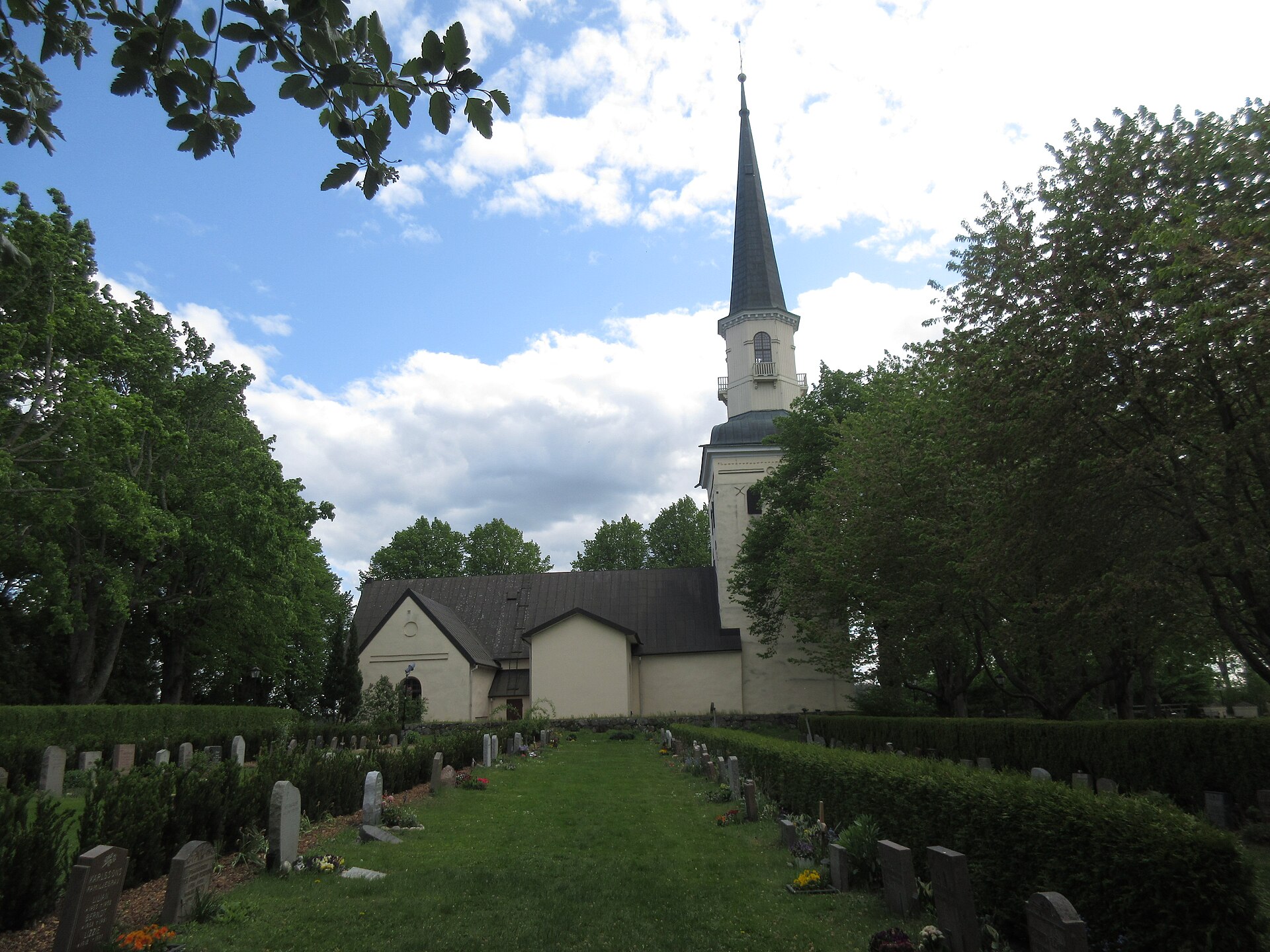 Ekerö kyrka i gul puts med högt torn och spira, sedd genom kyrkogården