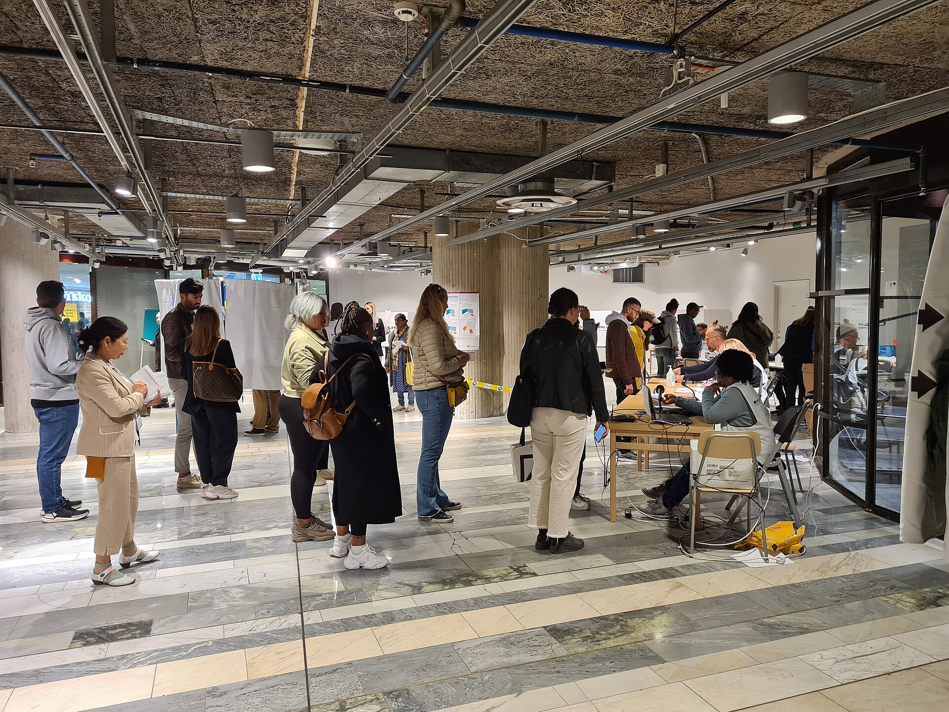 Väljare står i kö till röstmottagarnas bord i en lokal för förtidsröstning, med valskärmar i bakgrunden