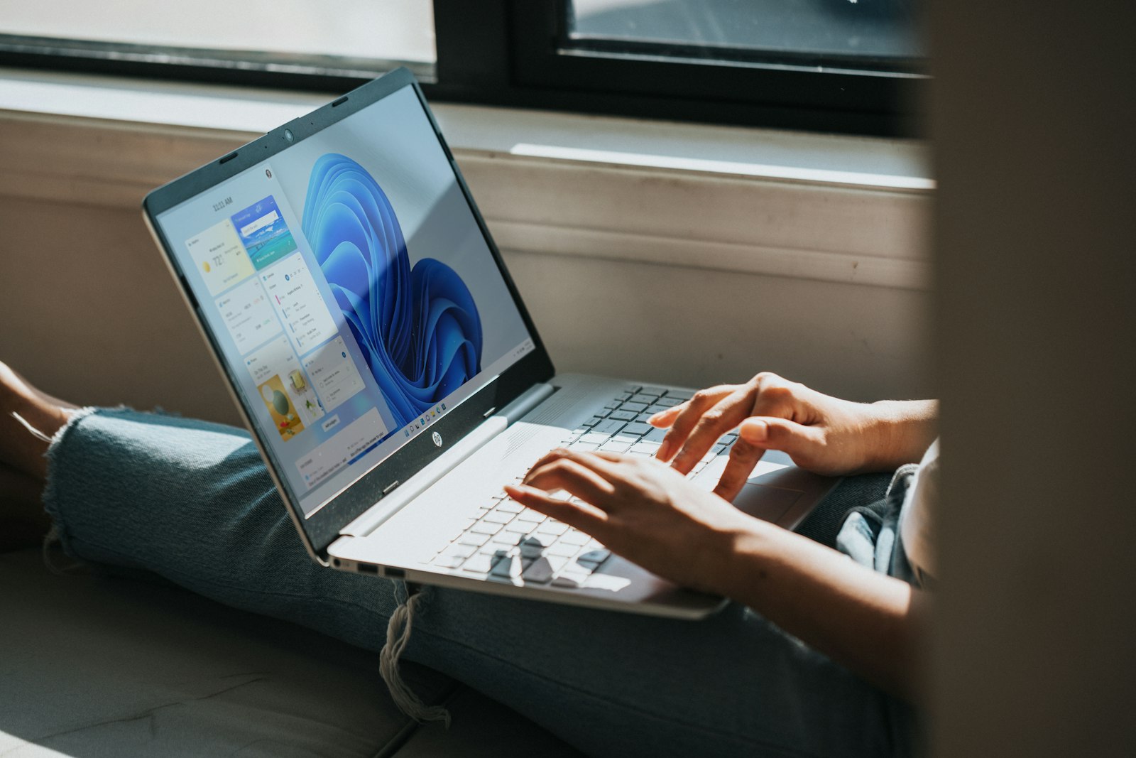 Händer som skriver på en bärbar dator i knät, där skärmen visar Windows 11-skrivbordet med blå bakgrund och widgetpanelen öppen.