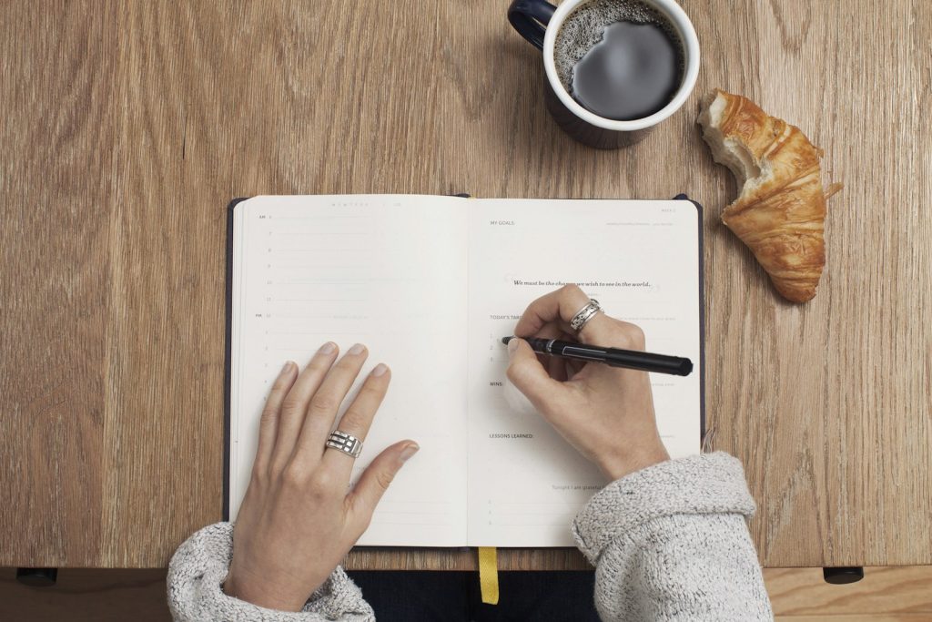 Person som skriver för hand i en anteckningsbok vid ett träbord med kaffe