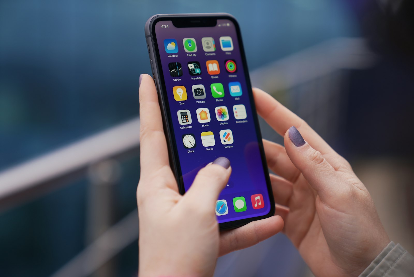 En hand håller upp en iPhone med hemskärmen synlig, med appikoner som Väder, Kamera, Kalkylator och Klocka i rader